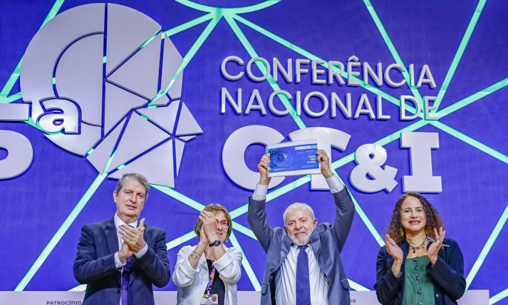 President Lula holds up the PBIA document on the stage of the 5th National Conference on Science, Technology and Innovation, applauded by three people